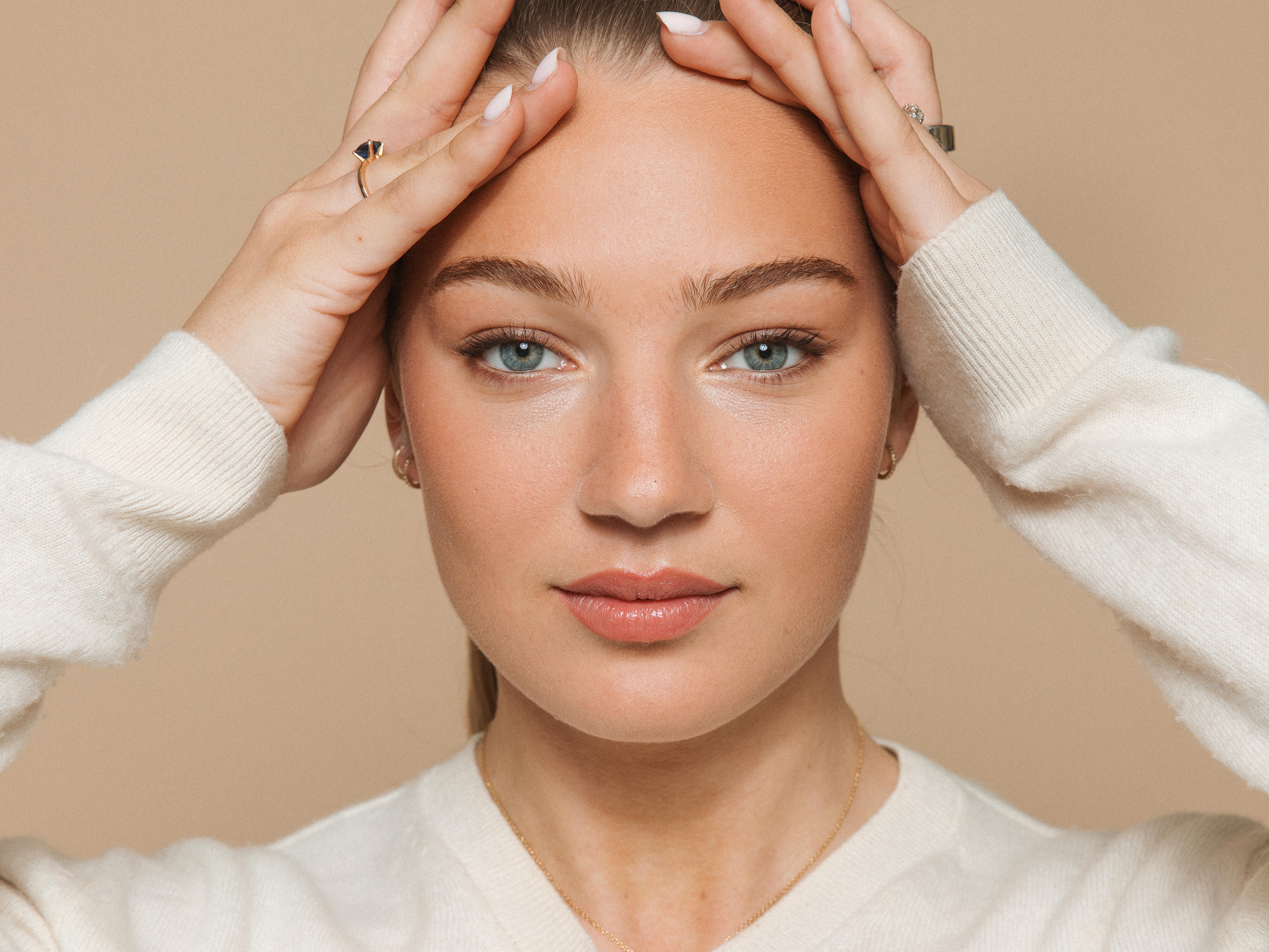 Soap Close-up portret van een vrouw met natuurlijke make-up en blauwe ogen, haar handen tegen haar voorhoofd houdend terwijl ze direct in de camera kijkt. Ze draagt een crèmekleurige trui en subtiele sieraden tegen een beige achtergrond.